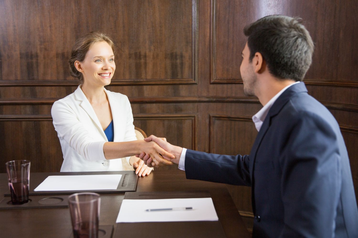 A professional in a white blazer shakes hands with a colleague in a navy blue suit at a dark wood conference table.