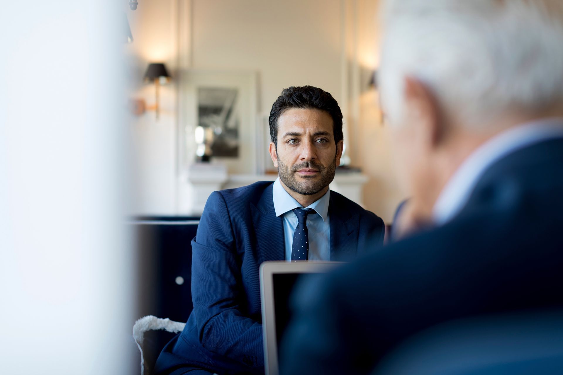 A professional in a suit and tie sits at a desk, looking intently at another person during a meeting.