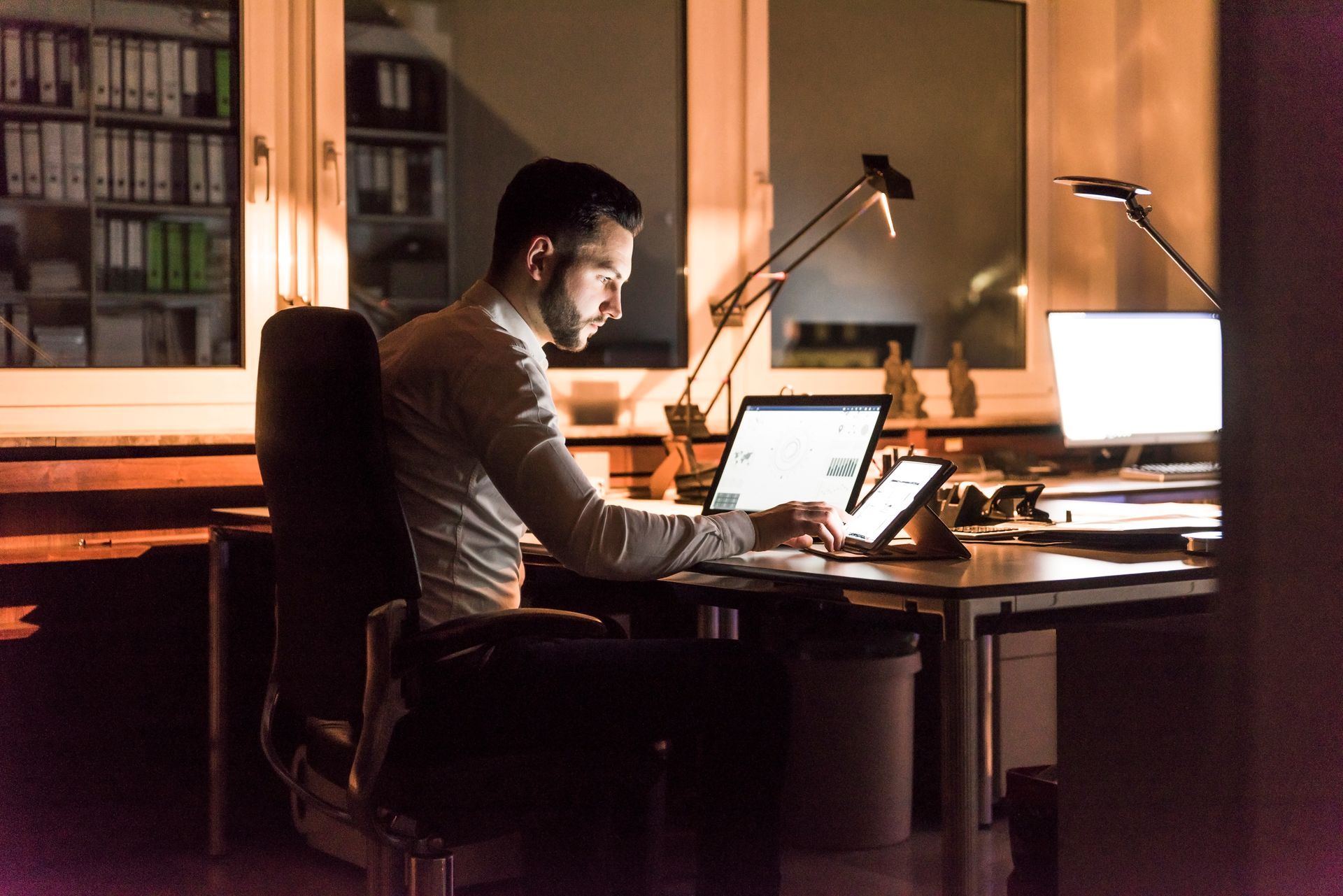 A person works late in a dimly lit office, using a laptop and tablet at a desk with an illuminated monitor.
