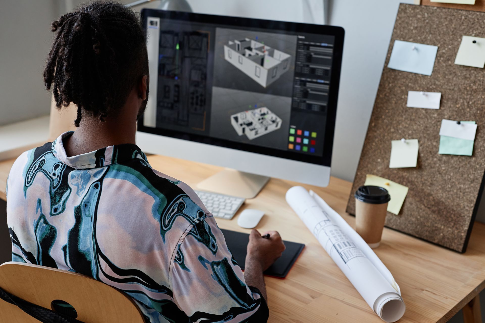 A person works at a desk, using a stylus to design a 3D building floor plan on a computer monitor.