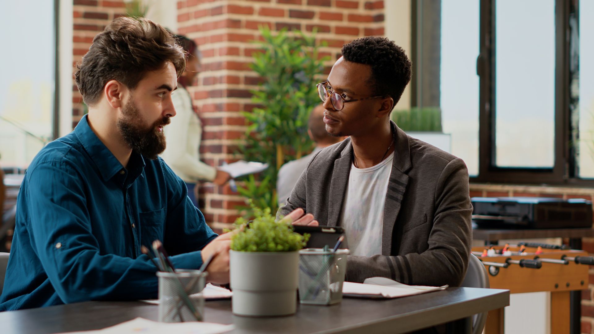 Two colleagues sit at a desk in an office, discussing work while looking at a tablet together.