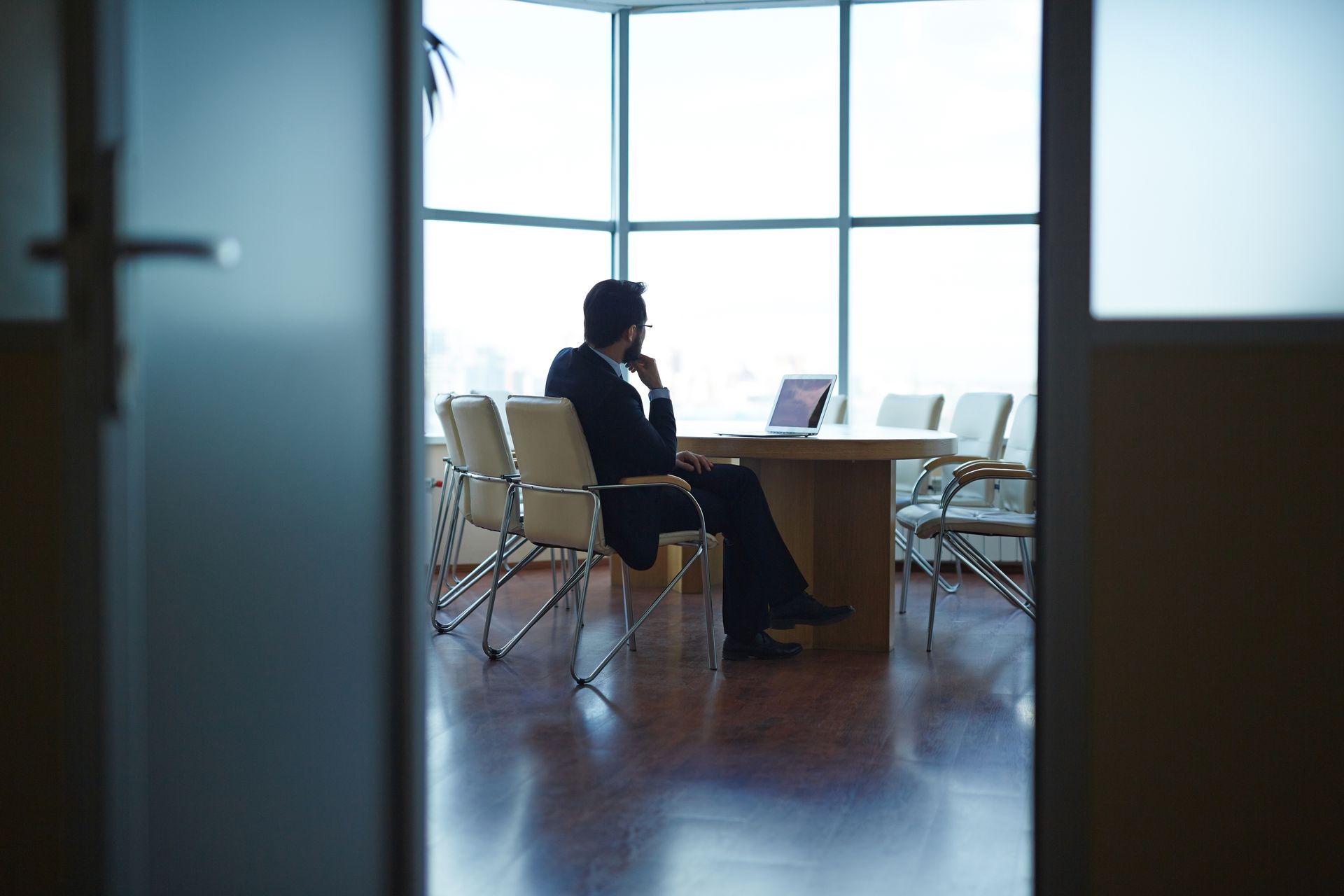 A person in a suit sits at a conference table, looking out of a large window in a bright office space.