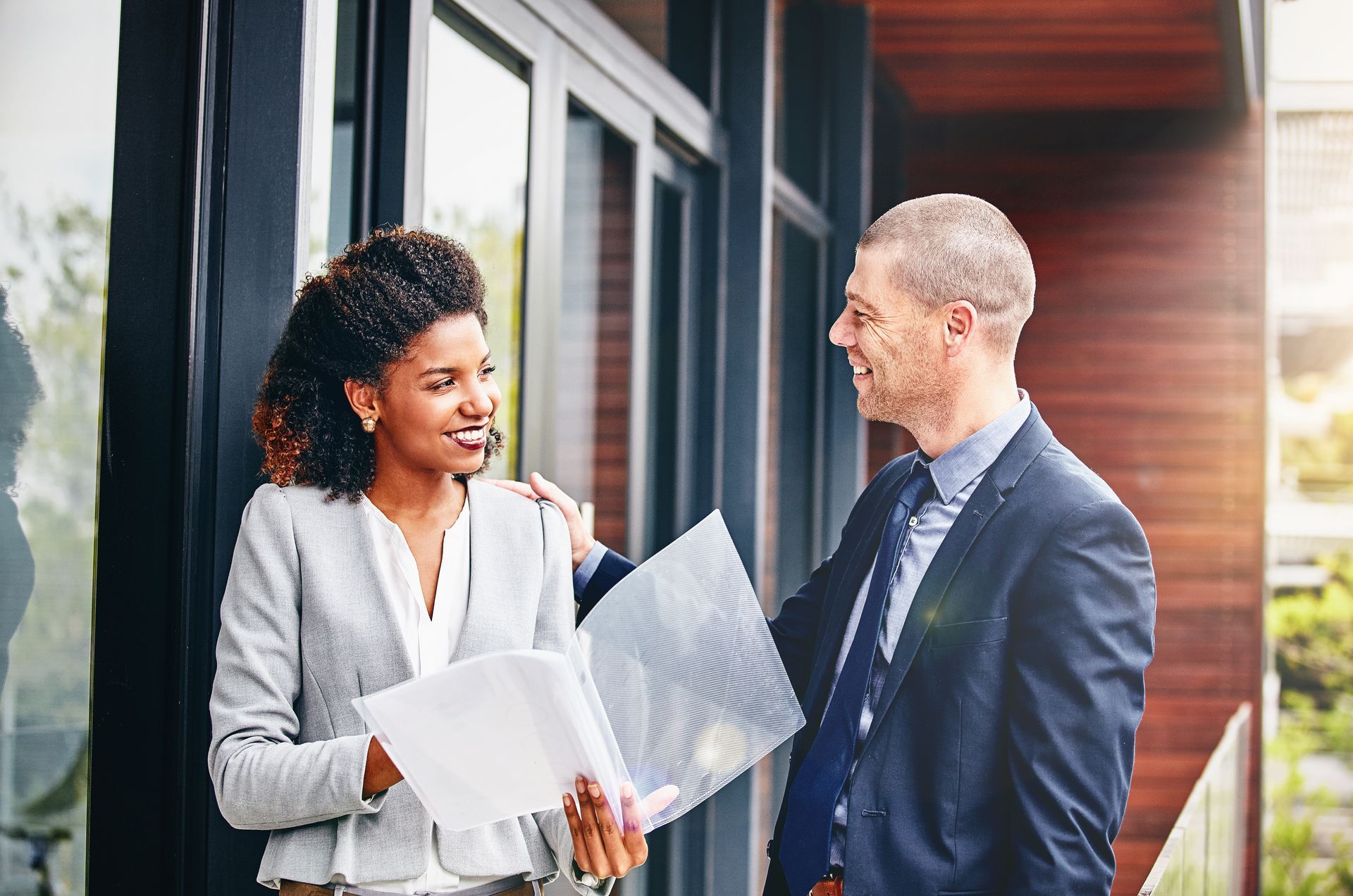 Two professionals in business attire hold a folder and converse while leaning against a modern building entrance.