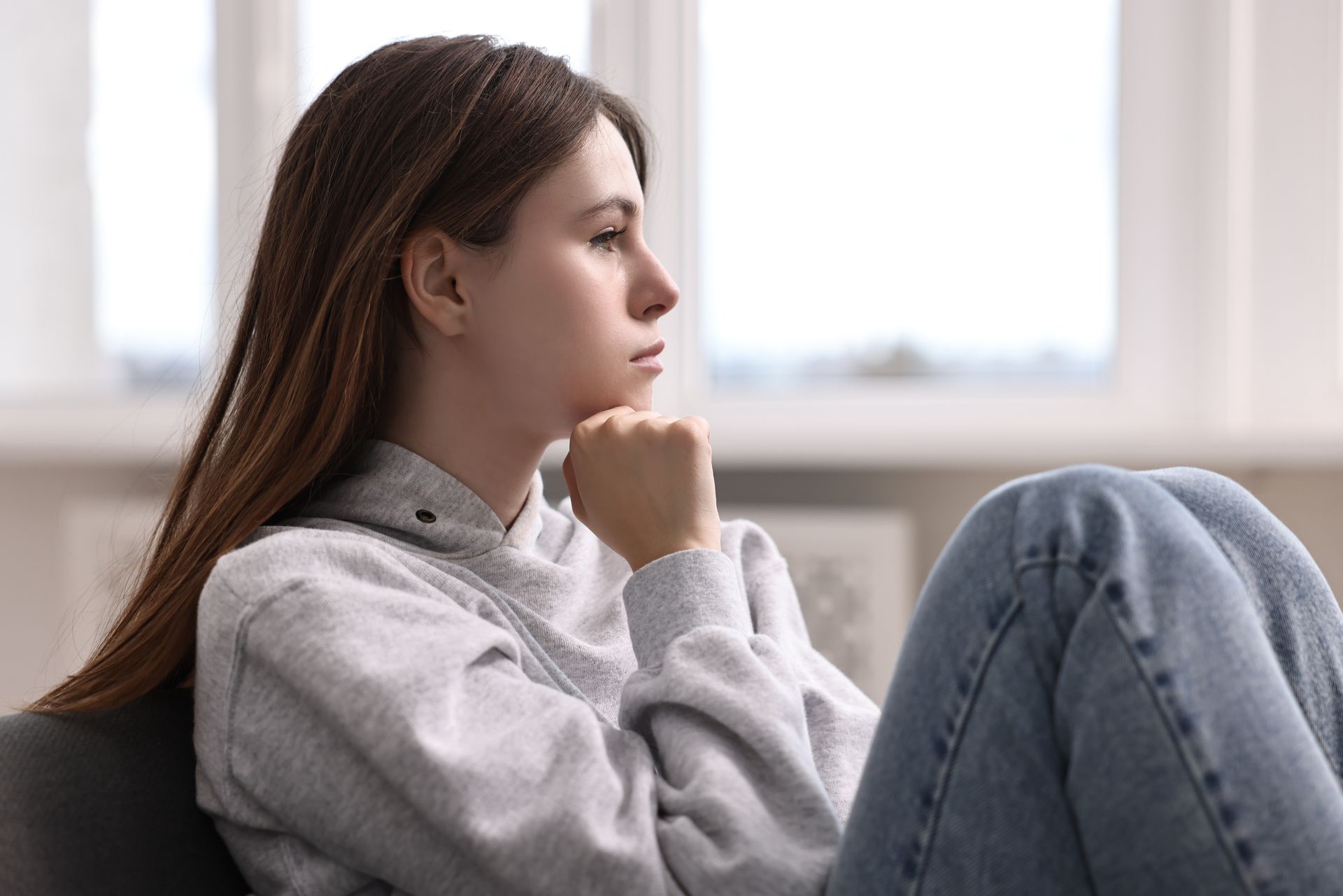 A person in a grey hoodie sits with their hand on their chin, looking thoughtfully toward a bright window.