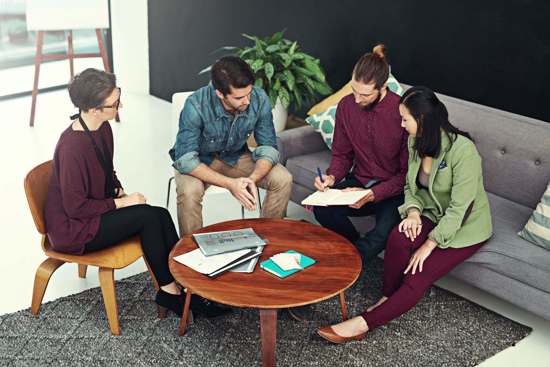 Four people sit in a casual office meeting around a wooden coffee table, engaged in a discussion with papers and a laptop.