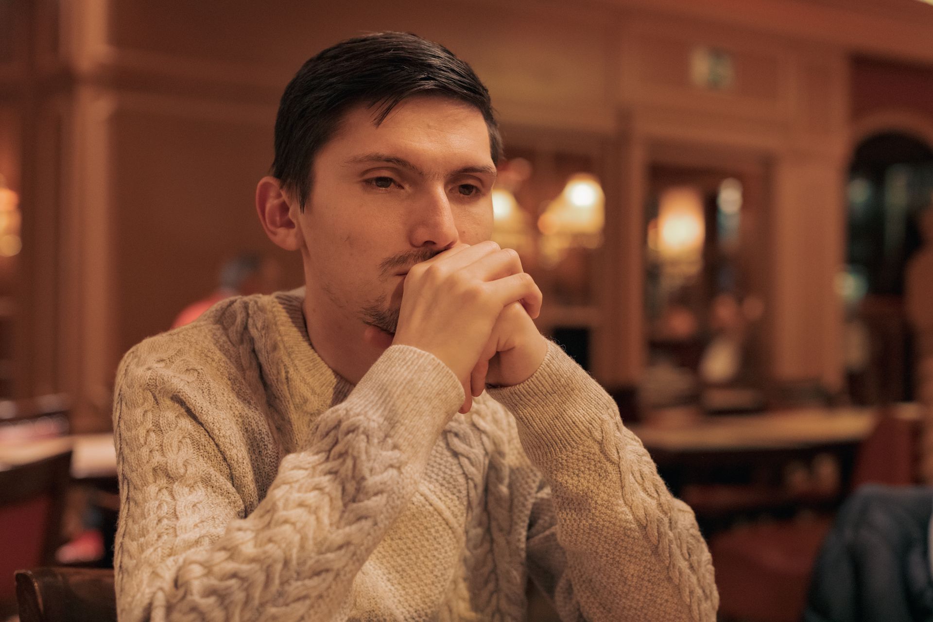A person in a cream cable-knit sweater sits in a warm-toned room, resting their chin on their clasped hands.