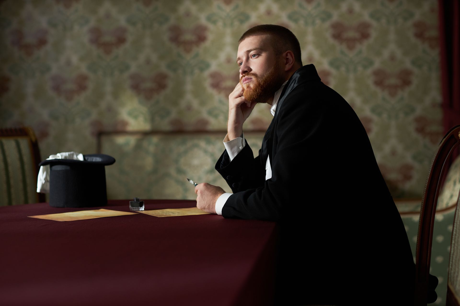 A thoughtful person with a beard sits at a maroon table with an ink pen, looking away beside a top hat in a patterned room.