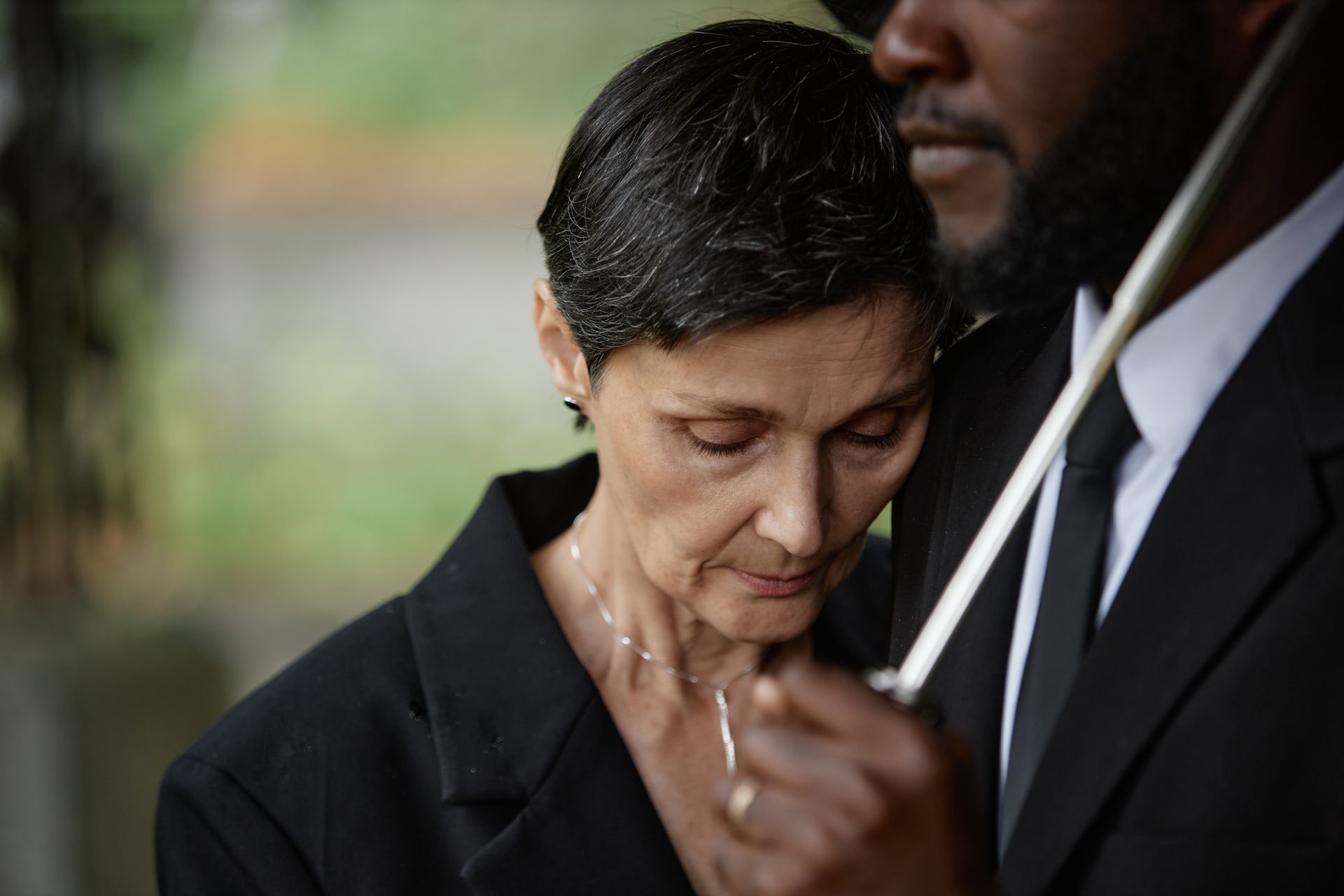 A person leans their head on another’s shoulder under an umbrella in a somber, outdoor setting.