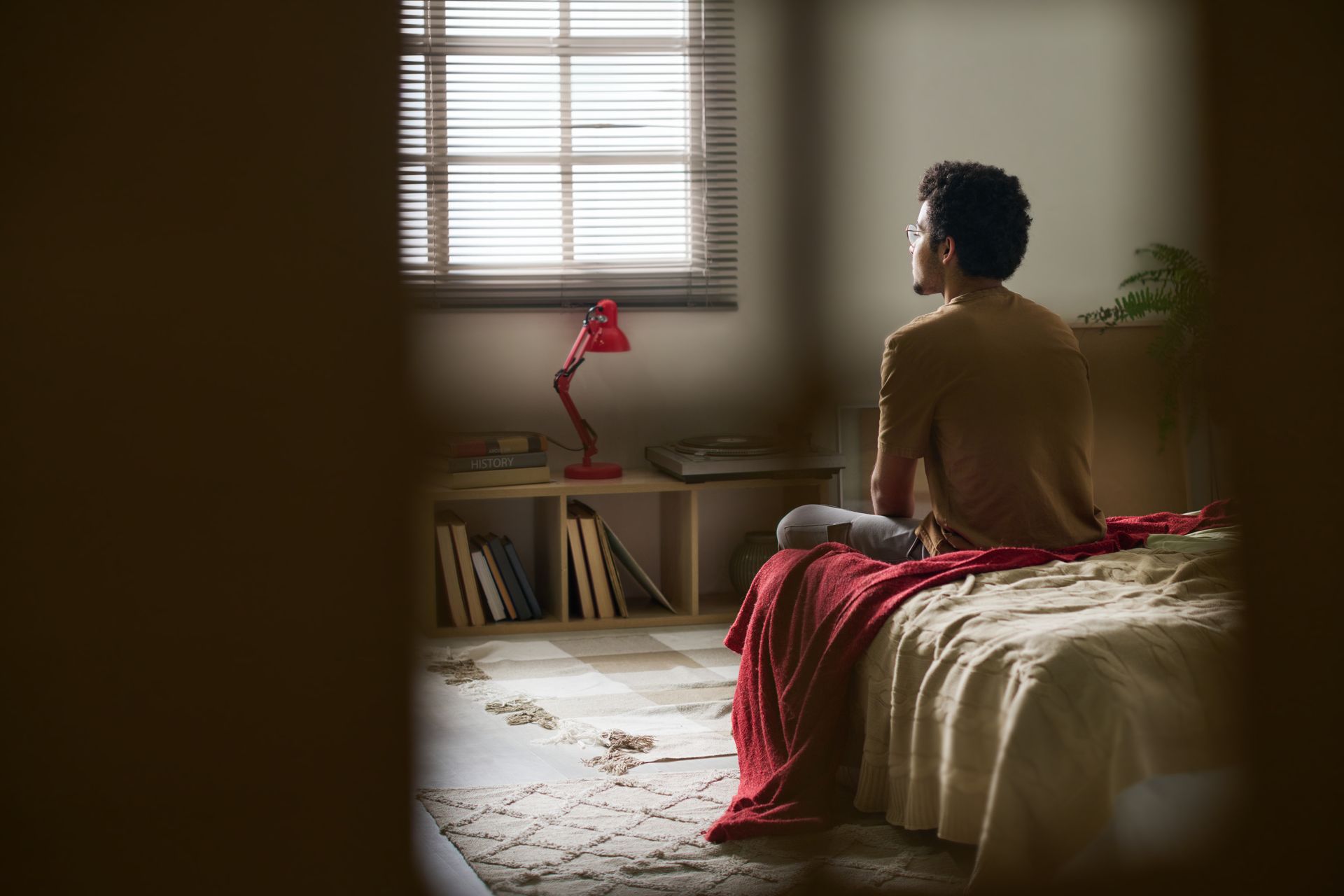 A person sits on a bed in a sunlit room, facing a window with blinds, with a red lamp and bookshelves in the background.