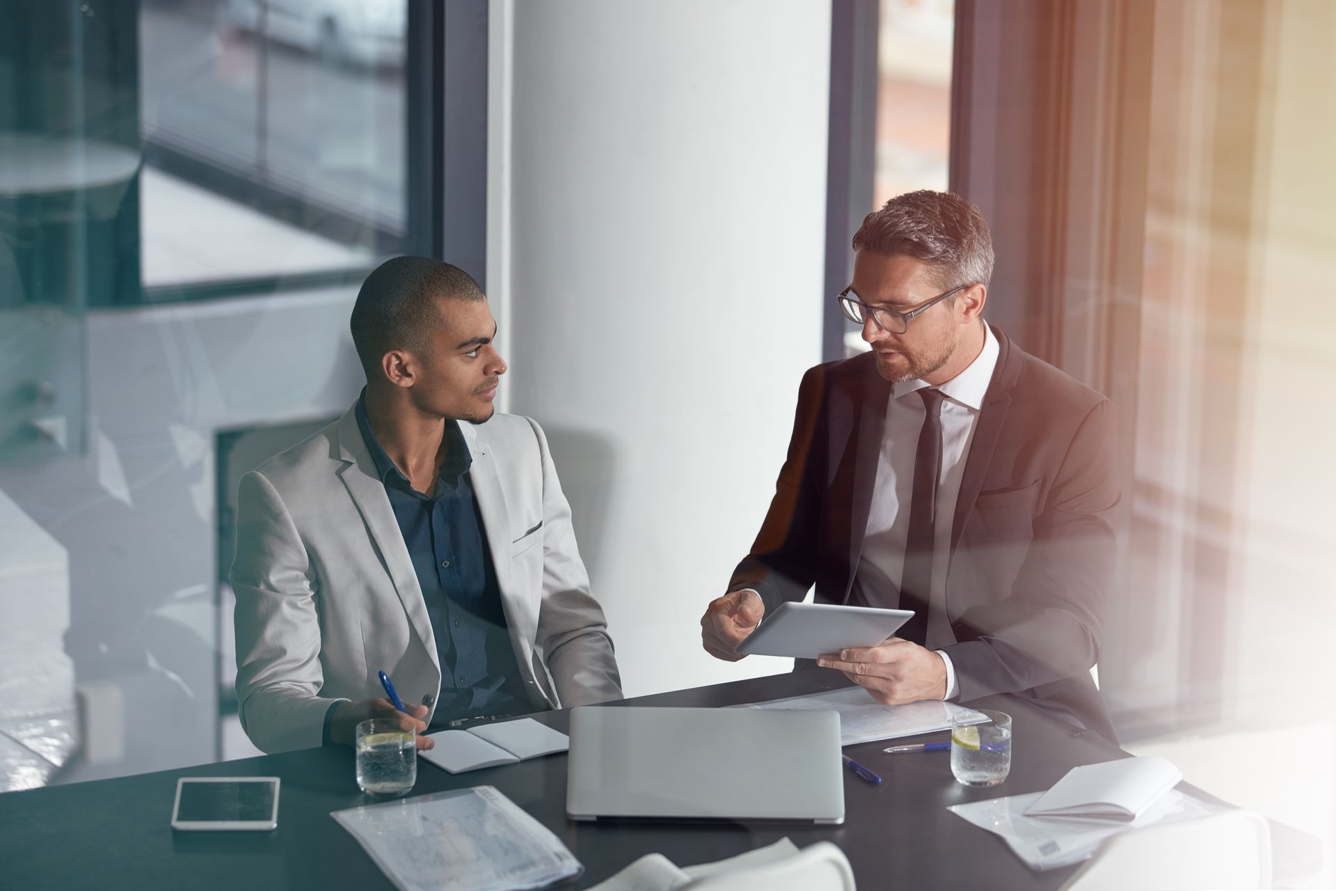 Two professionals in suits discuss work over laptops and documents in a modern office meeting room.