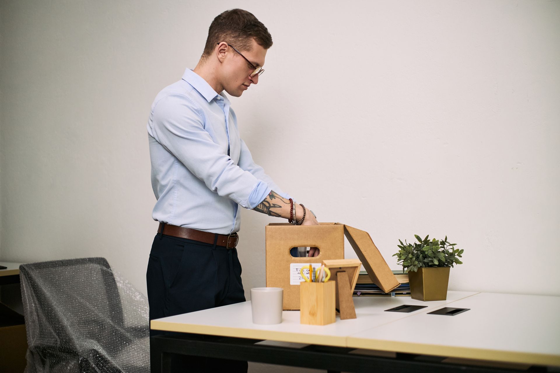 A person in a light blue shirt packs personal belongings into a cardboard box on a desk.