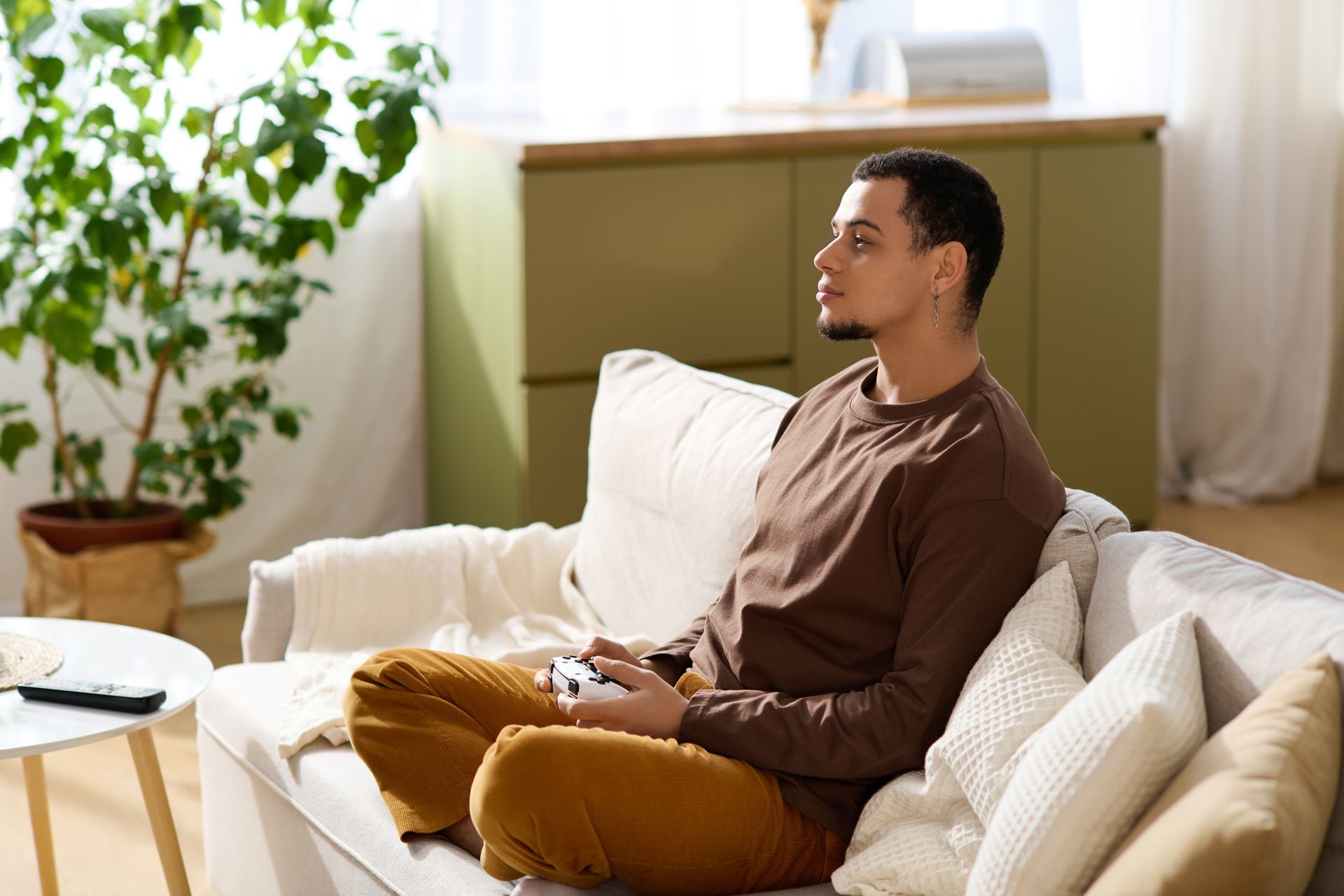 A person sitting on a sofa in a living room, holding a game controller and looking toward a screen.