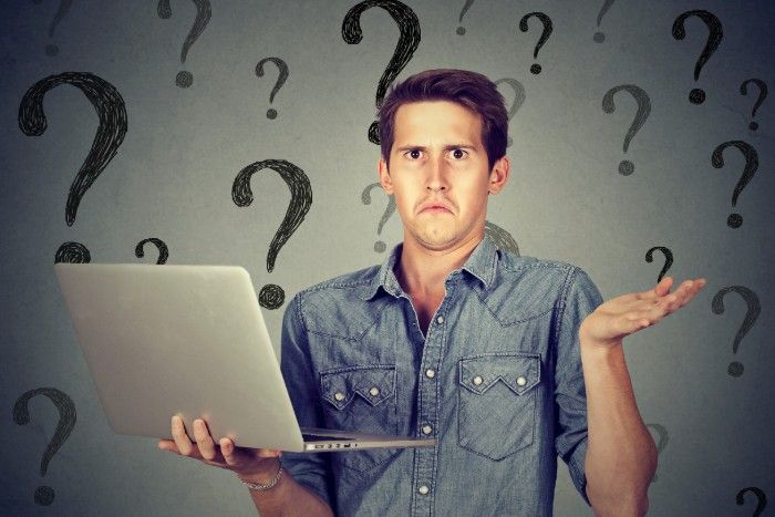 Man with laptop looks confused, shrugging, surrounded by question marks on a gray background.