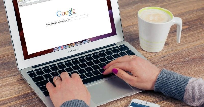 木製のテーブルの上に、Google 検索エンジンを開いたままノートパソコンでタイピングしている人、マグカップのコーヒー、スマートフォン。