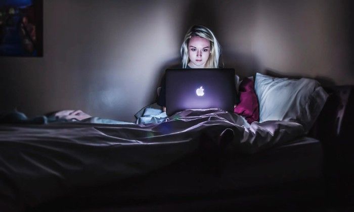 Woman with blonde hair illuminated by laptop screen in a dark bedroom, sitting on a bed.