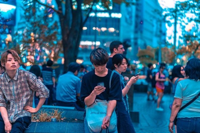 People on a city street at dusk; a man looks up, others on phones. Blue and green tones.