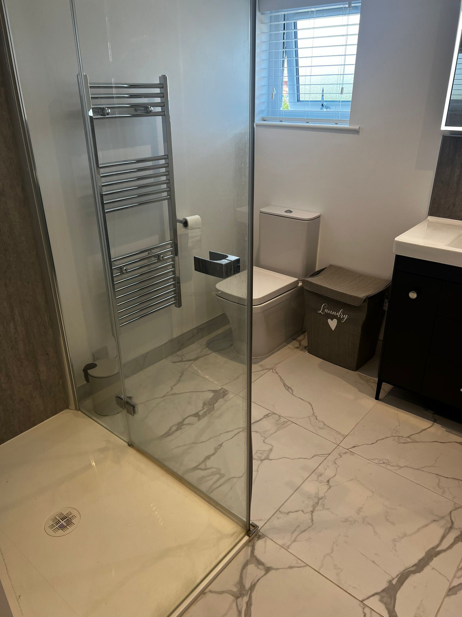 Bathroom with a glass shower enclosure, heated towel rack, toilet, and vanity. Marble-look tile floor.