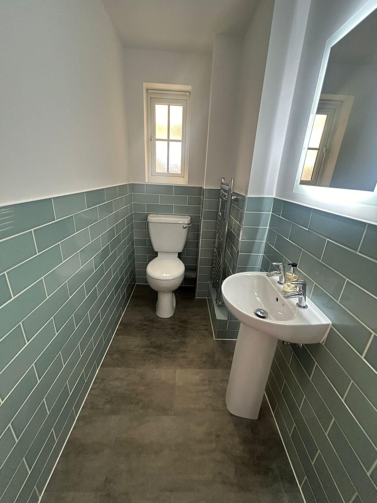 Small bathroom with blue-green tiled walls, white toilet and pedestal sink, small window, and grey floor.