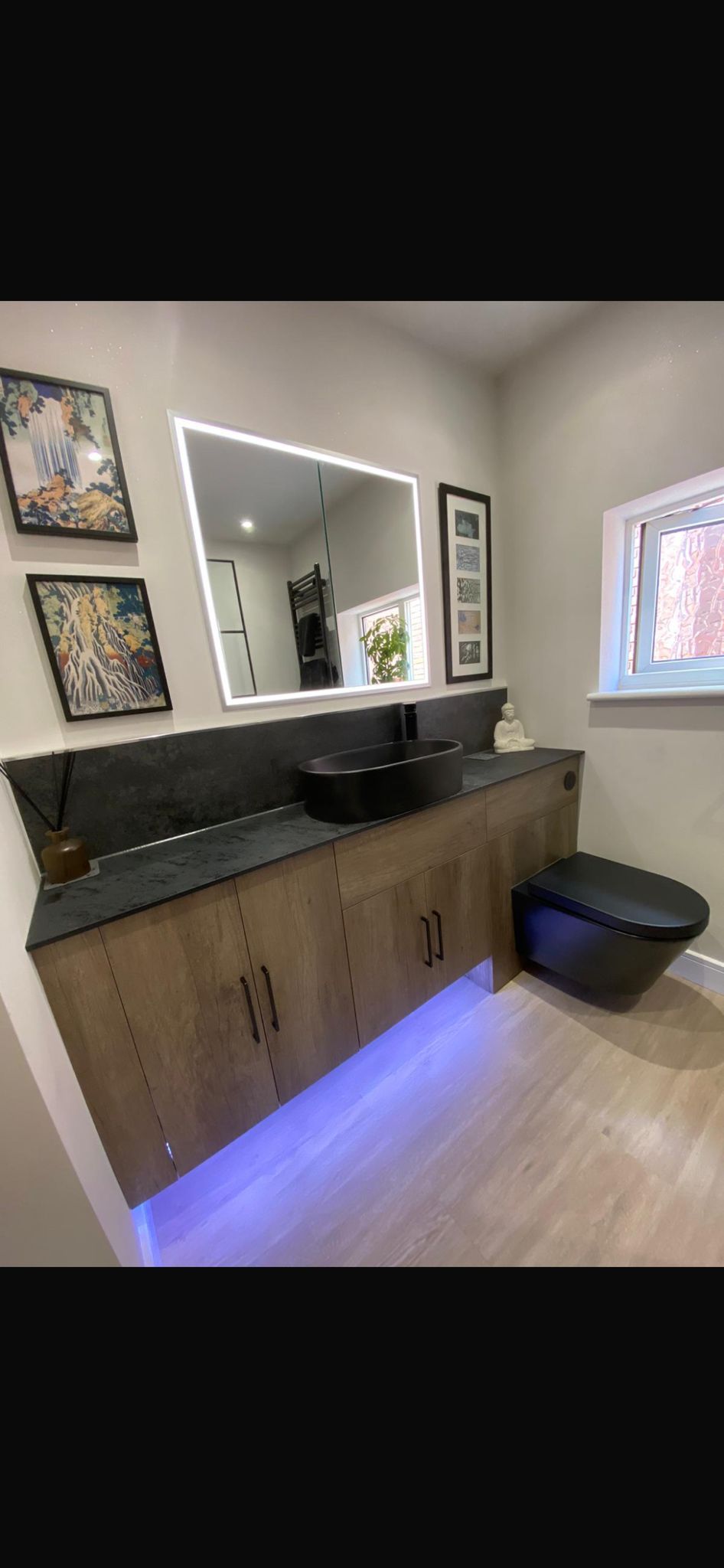 A modern bathroom with a wooden vanity, a black sink, and a backlit mirror. There are blue lights underneath the vanity and two framed pictures on the wall.