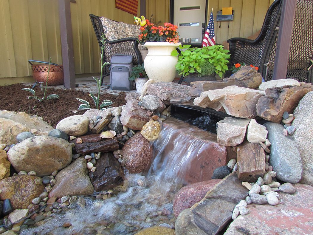Water cascading over rocks in a small garden pond, with plants and a seating area in the background.