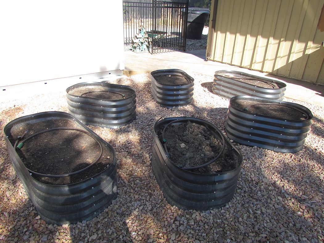 Six raised, galvanized metal garden beds filled with soil sit on gravel in sunlight.
