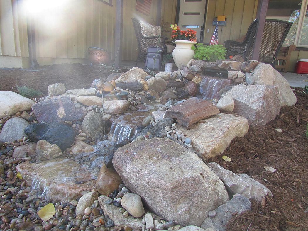 Water flows over rocks in a small garden, with a glimpse of a porch and potted plants in the background.