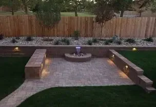 Backyard patio with a water feature, pavers, retaining walls, grass lawn, and a wooden fence.
