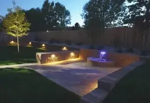 Nighttime backyard with illuminated fountain and retaining walls, trees, and pathway.