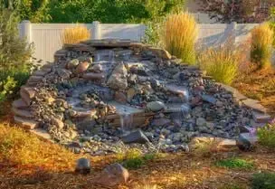 Waterfall feature in a yard with rocks and plants, wooden fence in the background.