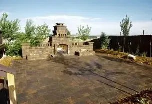Stone patio with a decorative archway and surrounding greenery under a blue sky.
