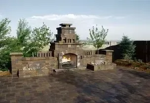 Stone outdoor fireplace with burning fire, brick patio, surrounded by trees and fence.