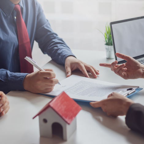 A picture of a man holding a pen over a contract with another person handing him the contract at a desk.