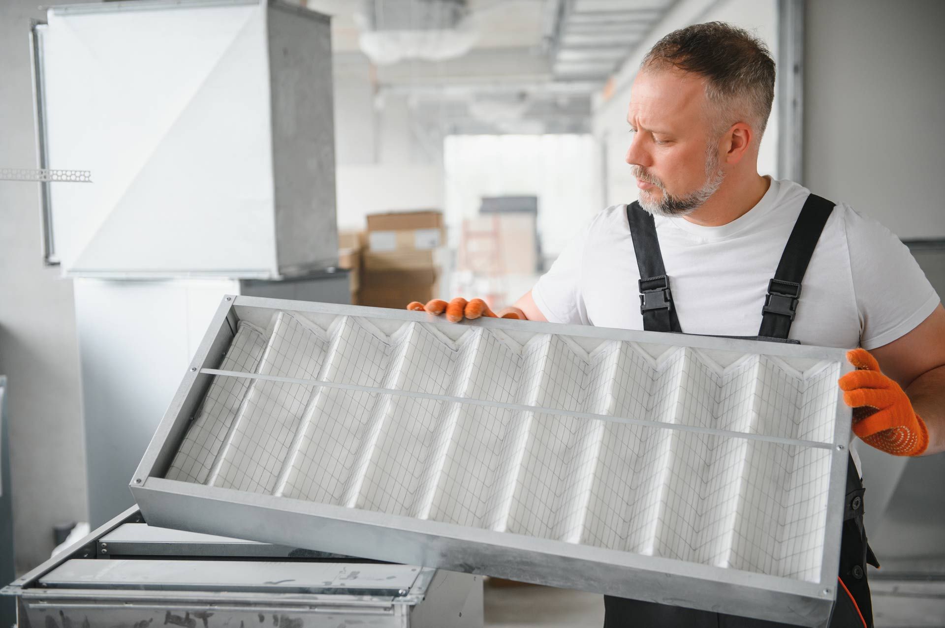 HVAC technician examining an air filter in a ductwork system, wearing work gloves and overalls.