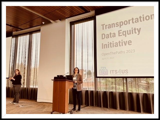 Dr. Anat Caspi presenting at a podium with screen behind with text that reads Transportation Data Equity Initiative Open The Paths 2023. To the left stands the sign language interpreter.