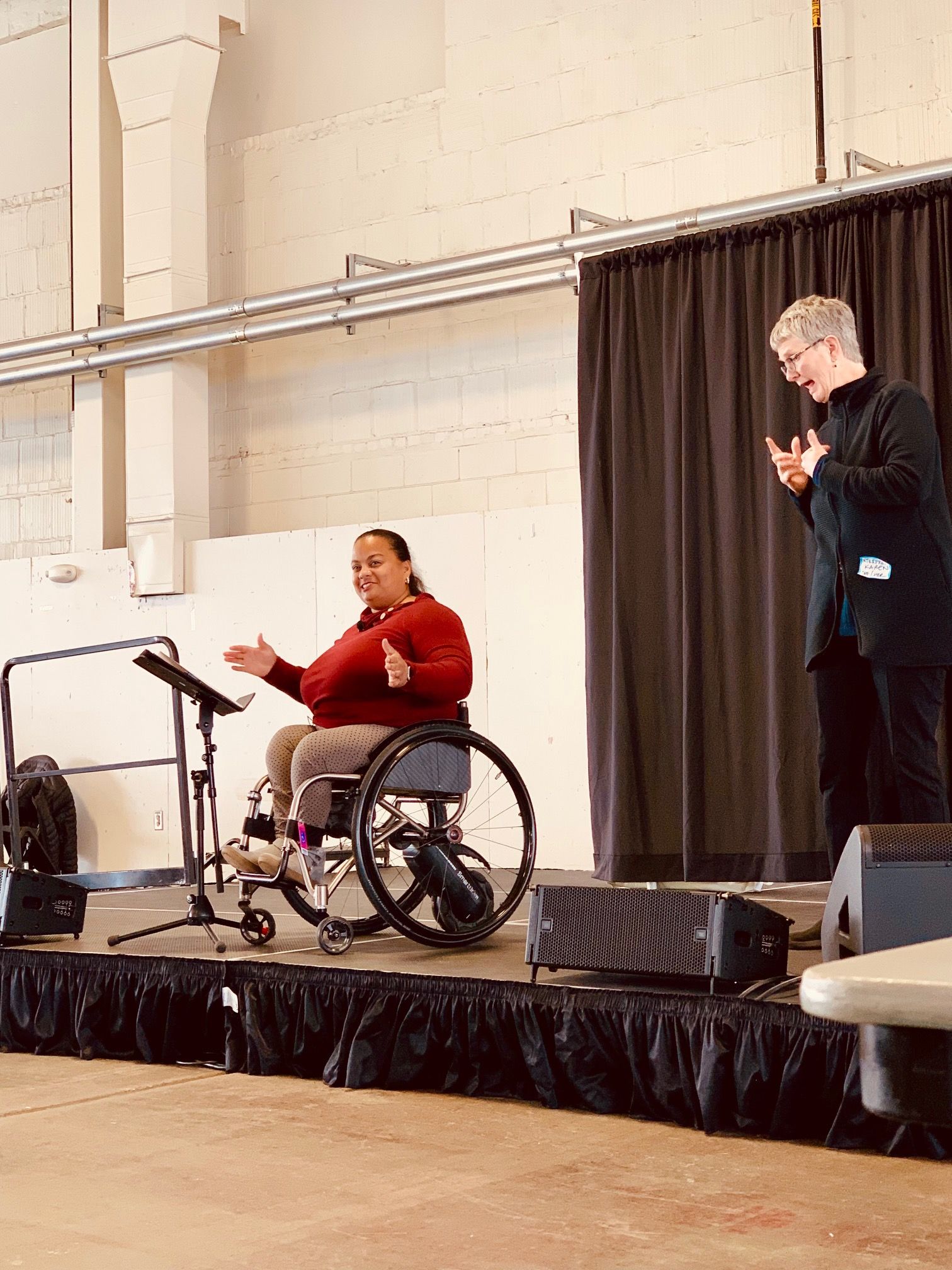 Anjali Forber-Pratt, PhD and the sign language interpreter on a stage. Dr. Forber-Pratt is wearing a red shirt and using her hands to talk about community and the Rehab Act