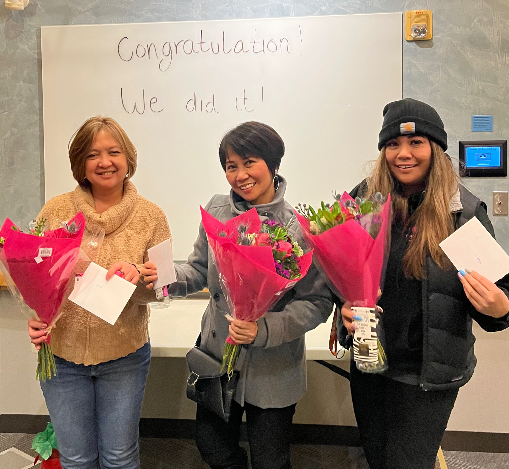 Three women holding flowers in front of a white board that says 