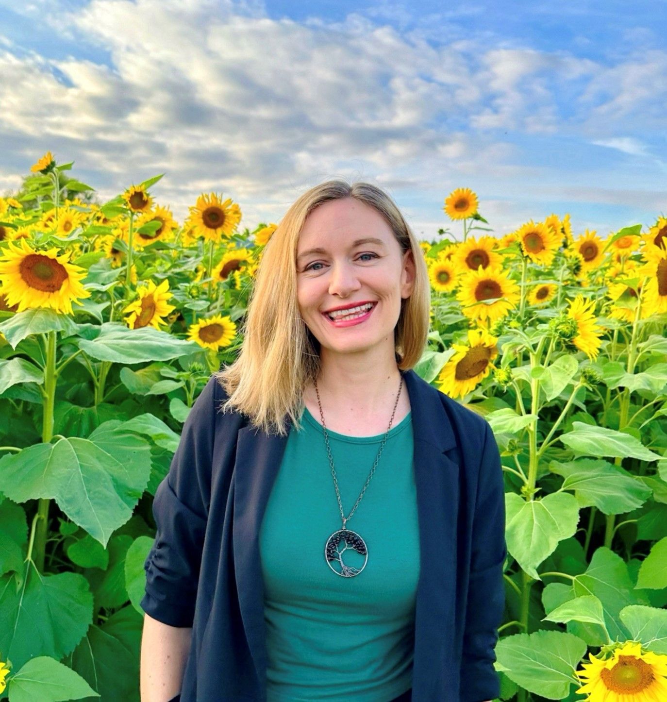 image of a white woman with dark, short hair. She is smiling at the camera and the background is a blooming shrub.