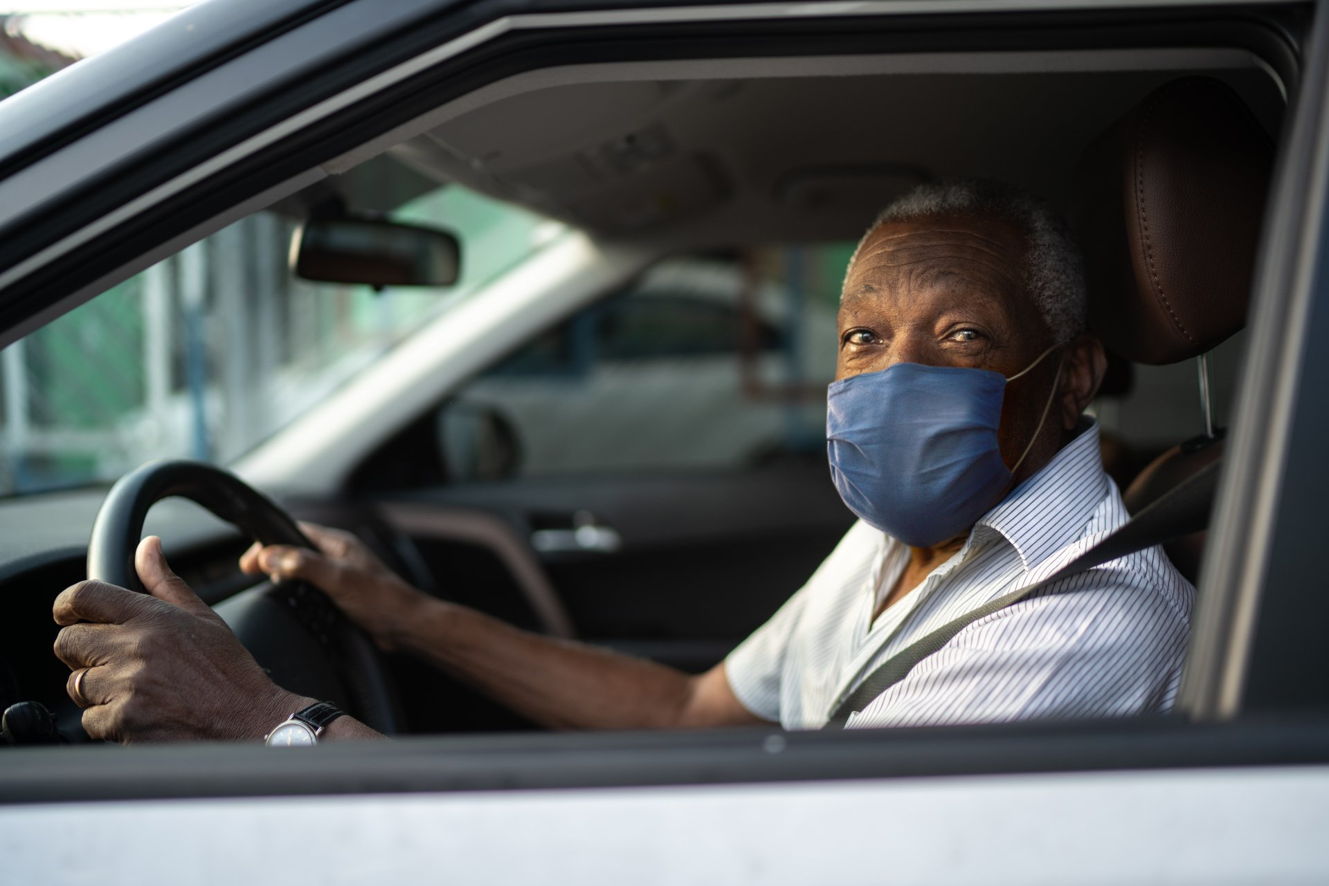 Older black man wearing a mask, in the driver seat of a car.