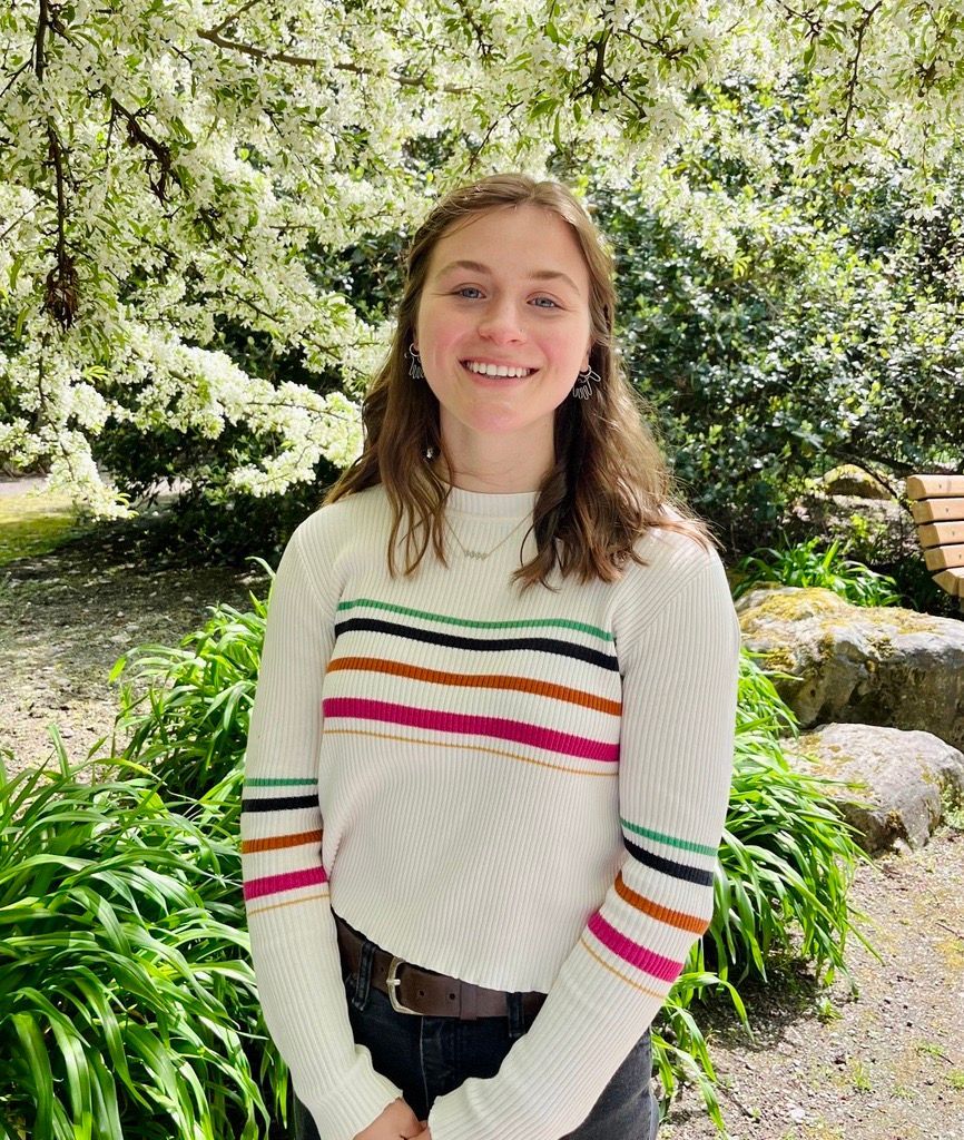 image of a white woman with dark, short hair. She is smiling at the camera and the background is a blooming shrub.