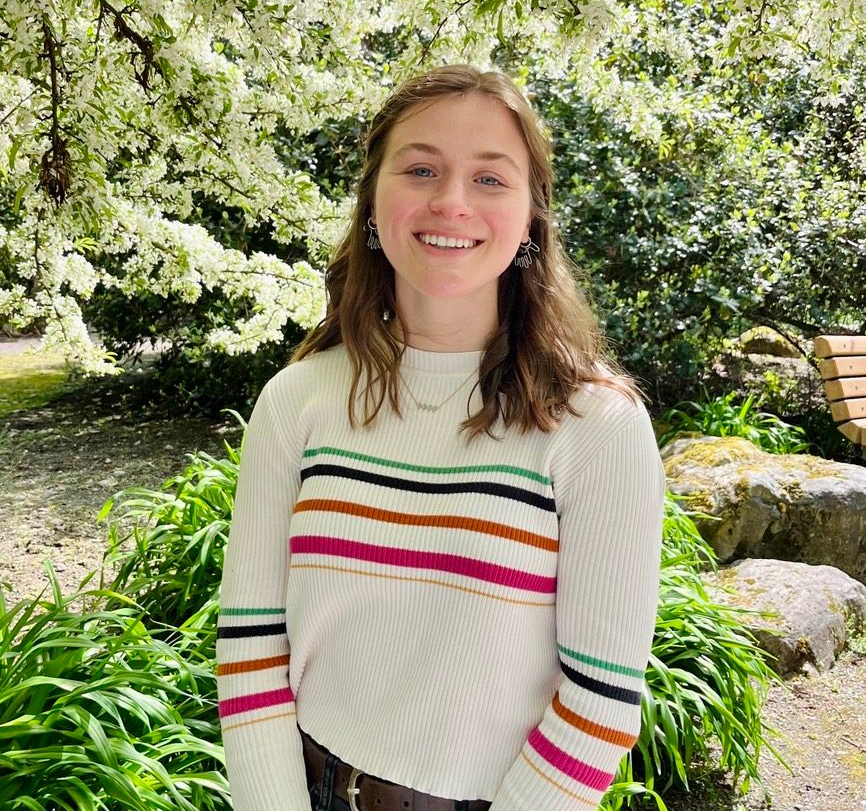 image of a white woman with dark, short hair. She is smiling at the camera and the background is a blooming shrub.