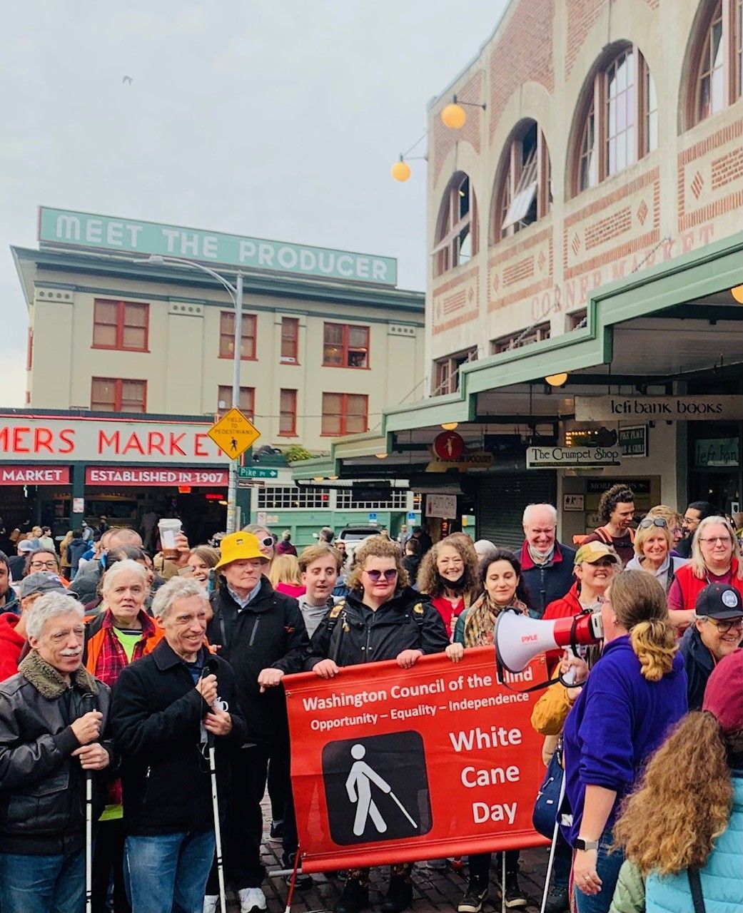 A group of people in front of Pike Place Market. A person with a megaphone speaking to people holding a sign and many attendees with white canes.