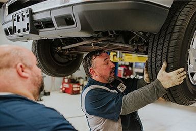 Two mechanics changing a tire under a vehicle in a garage. | Emge's Car Craft Auto Services