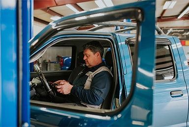 Mechanic in a blue truck, looking at a device in a garage. Door is open. | Emge's Car Craft Auto Services