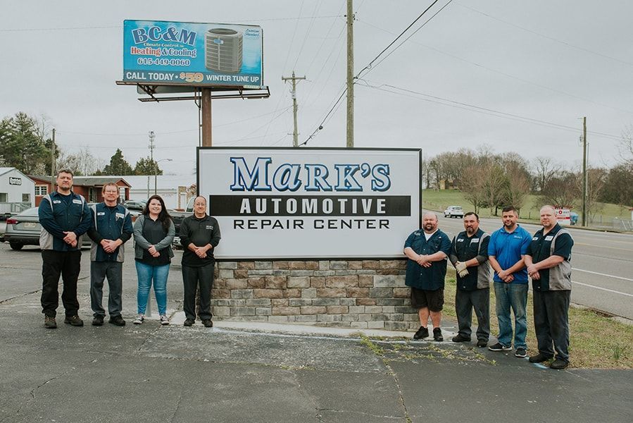 Group of people standing in front of Mark's Automotive sign. Cloudy sky, asphalt road, and buildings in the background. | Emge's Car Craft Auto Services