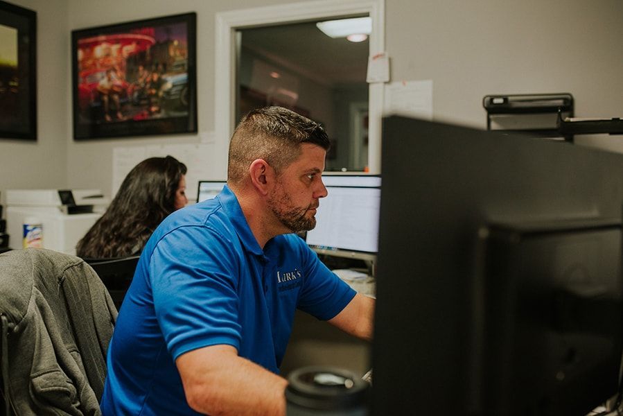 Man in blue shirt working at a computer in an office, with another person visible in the background. | Emge's Car Craft Auto Services