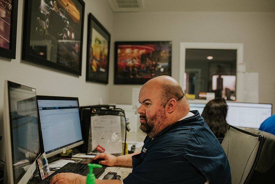 Man at desk, working on computer in an office with framed artwork on wall, another person in background. | Emge's Car Craft Auto Services