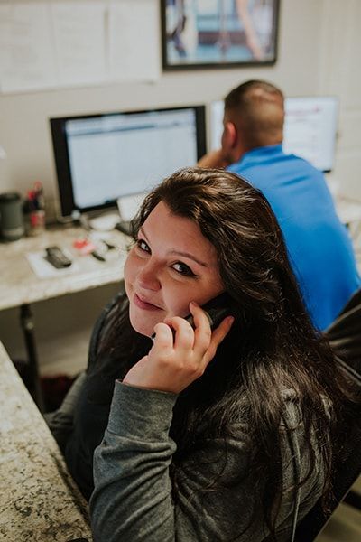 Woman on phone, smiling at camera, desk with computer in background. Man at second computer. | Emge's Car Craft Auto Services