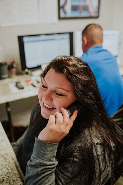 Woman smiling while talking on the phone in an office, with a man blurred in the background. | Emge's Car Craft Auto Services