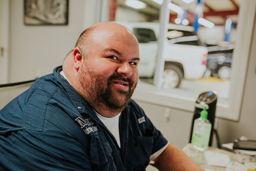 Man in blue shirt smiles indoors, white truck visible through window. | Emge's Car Craft Auto Services