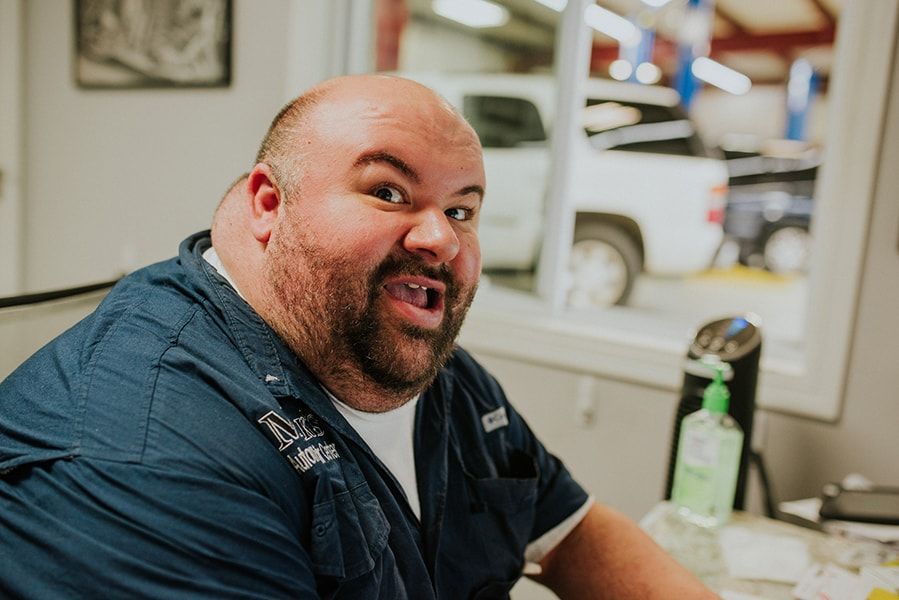 Man at desk in auto shop; looking at camera with open mouth, smiling; blue shirt. | Emge's Car Craft Auto Services